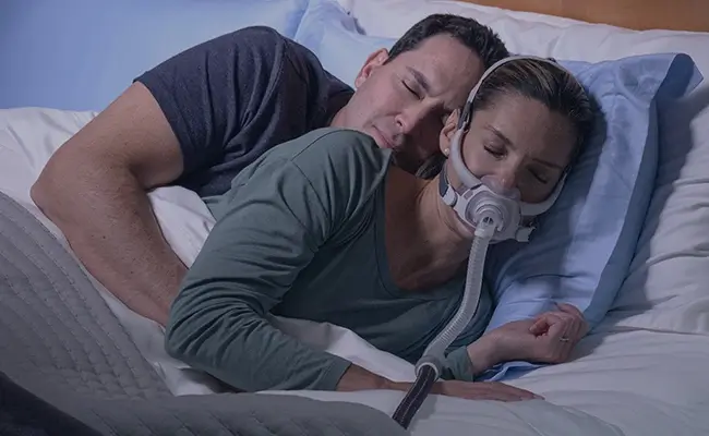 Et par som sover tett sammen i sengen, og der kvinnen bruker en AirFit F40 CPAP-maske.