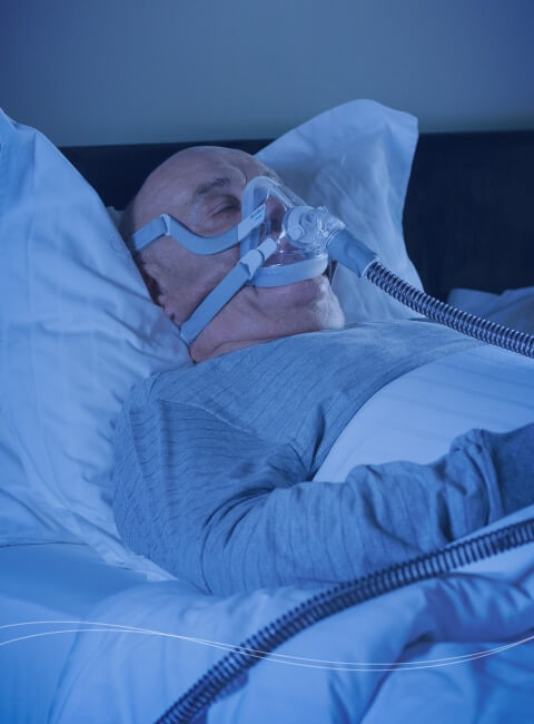 En mann som sover i sengen mens han puster gjennom en ventilator.