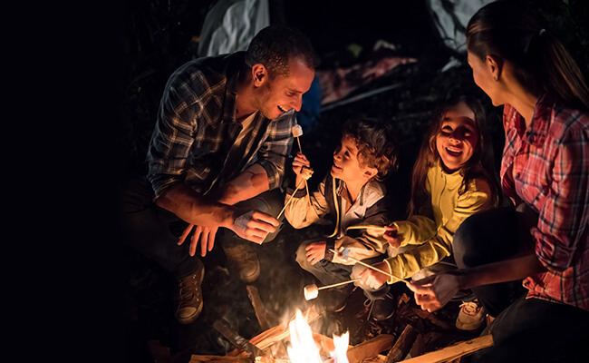 En familie med far, mor og to barn som griller marshmallows over en peis om kvelden