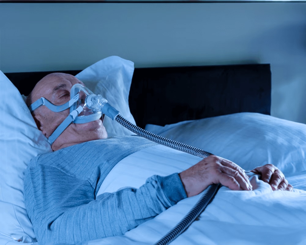 En mann som sover i sengen mens han puster gjennom en ventilator.